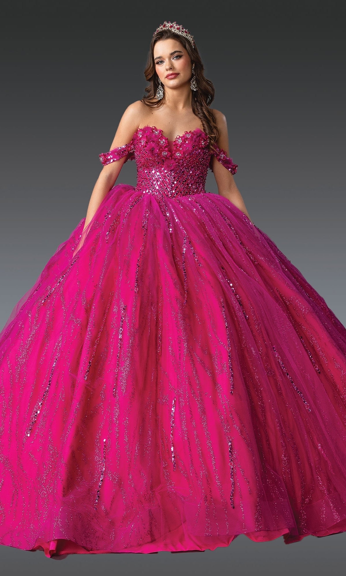 A woman in the Hot Pink Quinceanera Dress 1957 By Dancing Queen, featuring an off-the-shoulder design, glitter details, and a corset bodice with a matching tiara, stands against a plain gray background.